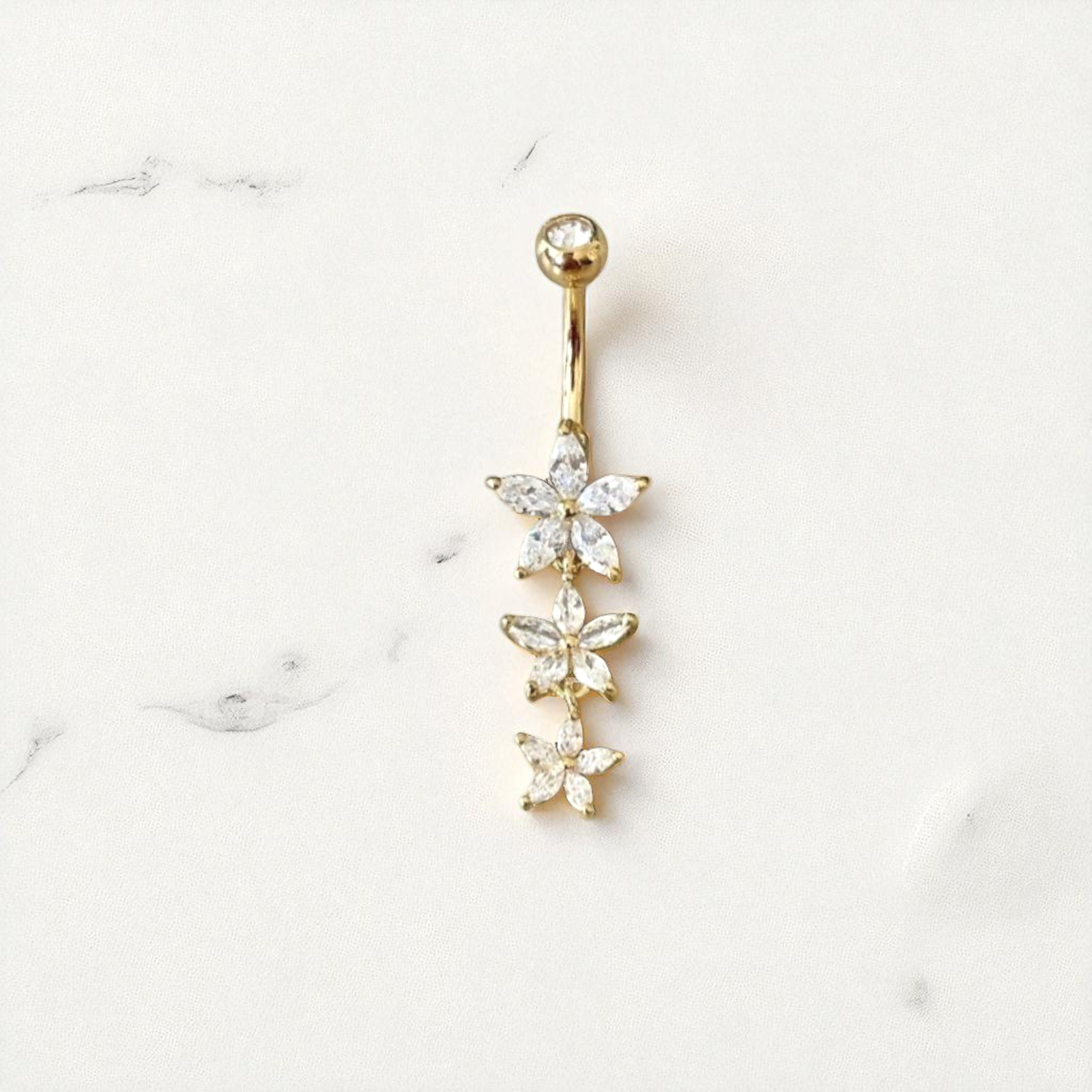 Gold belly button ring with flower-shaped gemstones on a white marble background