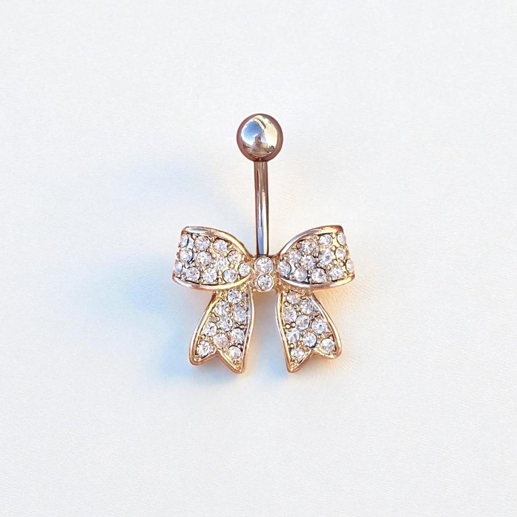 Gold bow-shaped belly button ring with clear gemstones on a light gray background