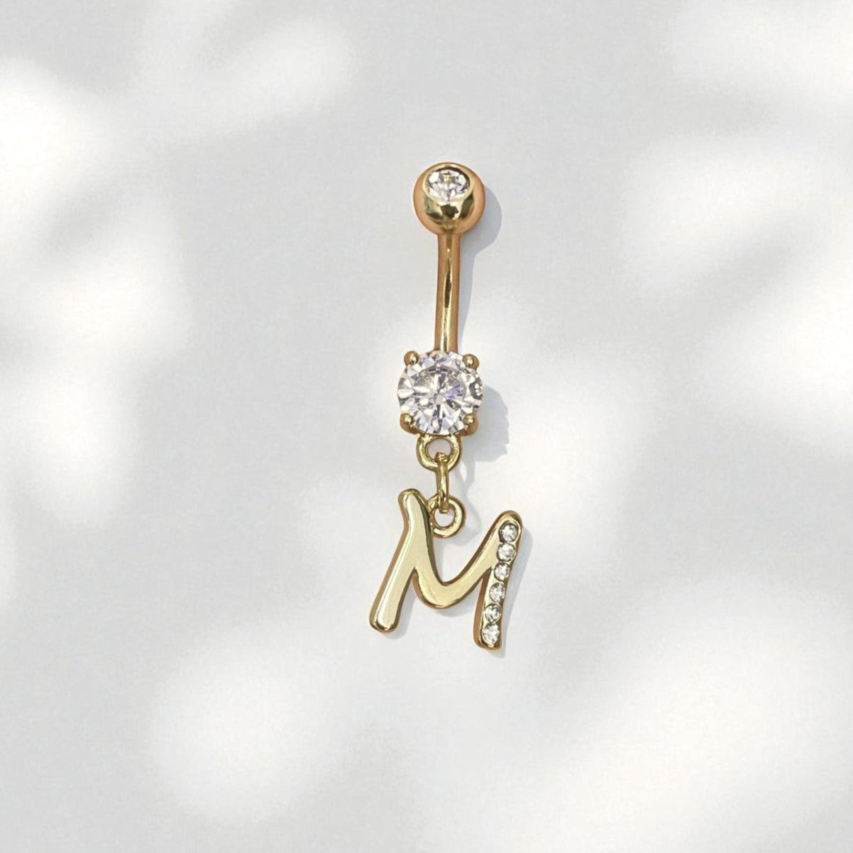 Gold belly button ring with 'M' pendant on a light gray background