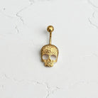 Gold skull-shaped belly button ring on a white cracked surface