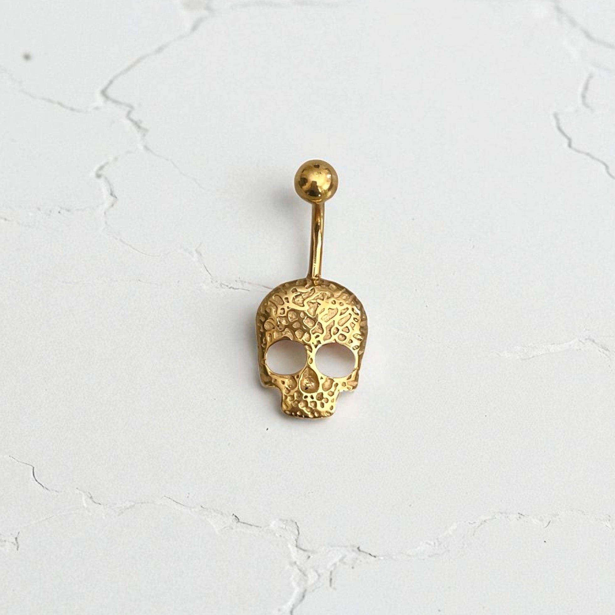 Gold skull-shaped belly button ring on a white cracked surface